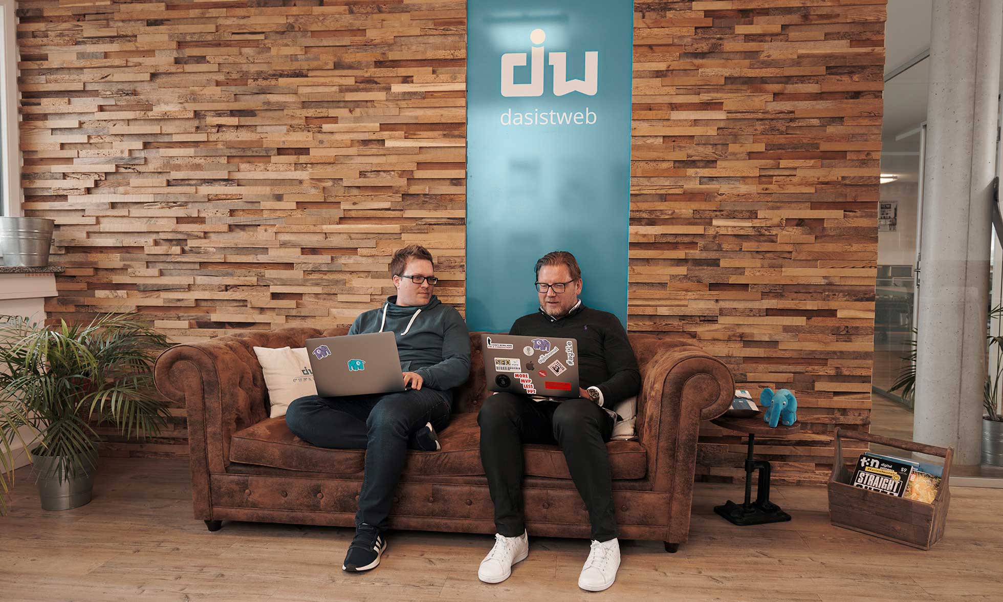 Martin Weinmayr and Andreas Rieger‚ CEO and Head of Sales at dasistweb sit on a sofa in their office. They are holding a laptop.